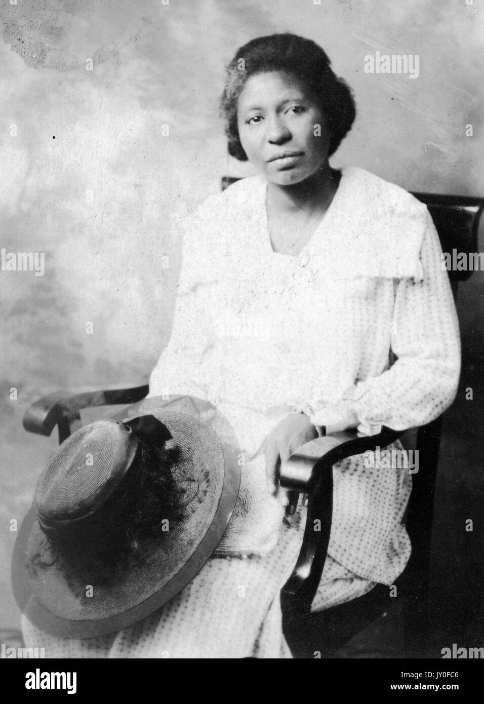 Dreiviertel sitzendes Porträt einer afroamerikanischen Frau mit neutralem Ausdruck vor einer gemalten Kulisse, mit Hut auf dem Schoß und einem leicht farbigen Kleid, 1920. Stockfoto