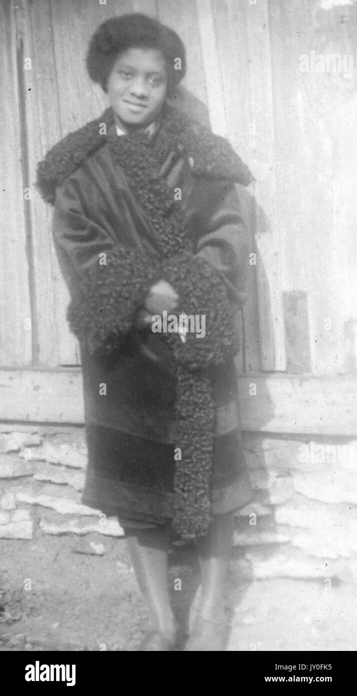 Volle Länge ständigen Porträt der jungen Afrikanische amerikanische Frau mit glücklichen Ausdruck, draußen vor dem Gebäude, das Tragen von langen, dunklen aufwendige Mantel, 1920. Stockfoto
