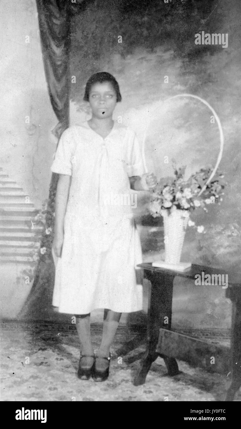 Ganzjähriges Standbild einer jungen afroamerikanischen Frau mit neutralem Ausdruck vor einer gemalten Kulisse, die ein leicht gefärbtes Kleid trägt und ihre linke Hand auf einem Blumenstrauß auf einem Tisch ruht, 1925. Stockfoto