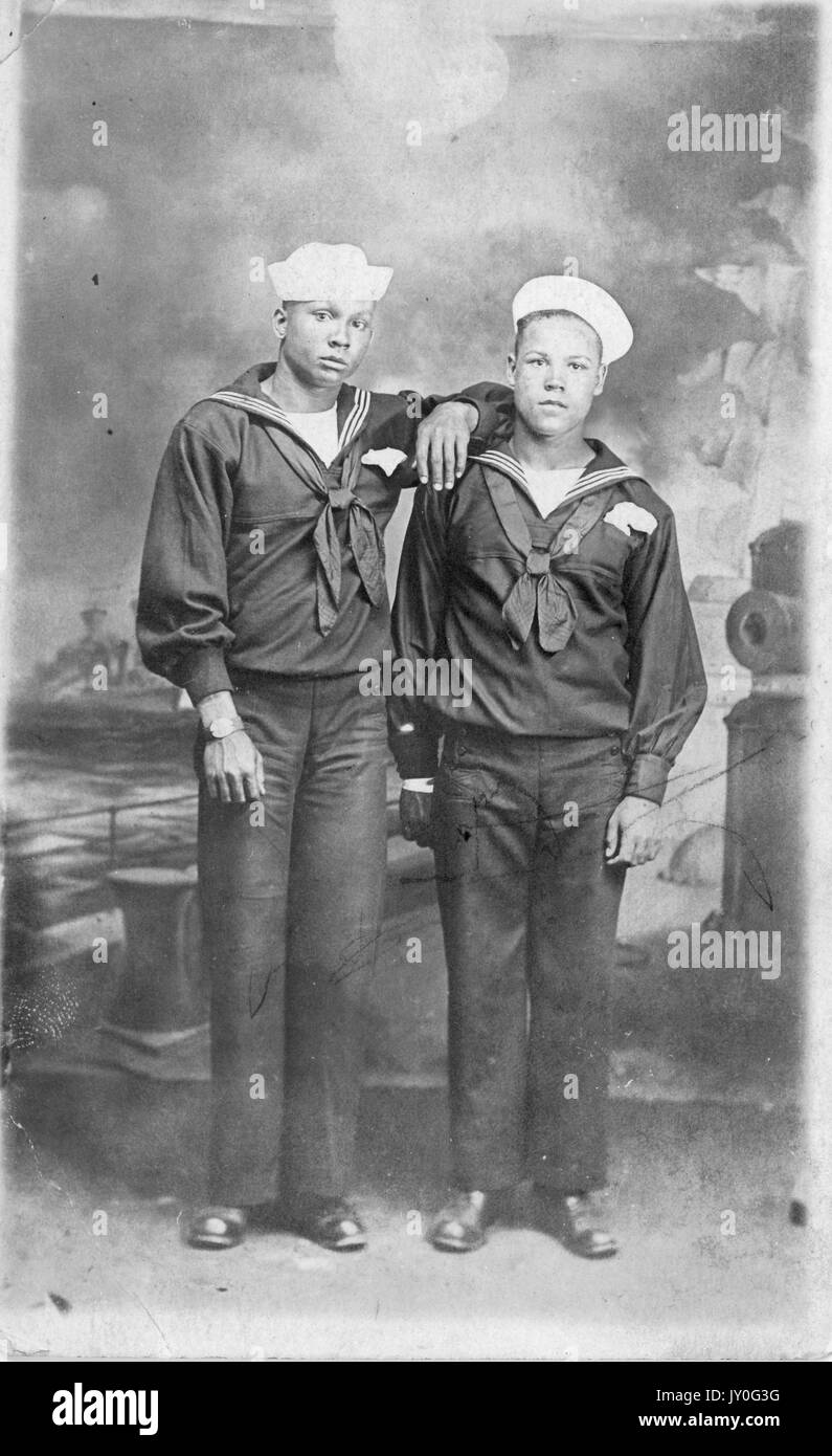 In voller Länge stehend Porträt von zwei jungen afroamerikanischen Männern mit neutralen Ausdrücken vor Hintergrund gemalt mit einer Hafenszene, in Matrosenkleidung gekleidet, 1920. Stockfoto