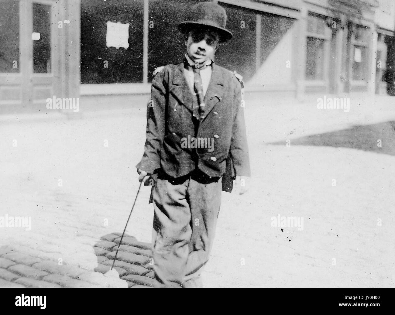 In voller Länge stehend Porträt von afroamerikanischen Kind, verkleidet als Charlie Chaplin in übergroßen Mantel und Hose, Krawatte und Melone Hut, tragende Stock, im Freien stehend, neutraler Ausdruck, 1925. Stockfoto