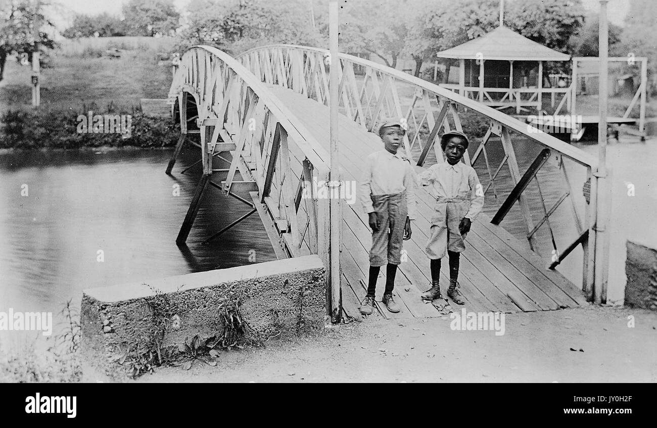 Langes Standbild zweier afroamerikanischer Jungen, in hellen Hemden, dunklen Hosen und Mützen, auf Brücke stehend, neutrale Ausdrücke, 1920. Stockfoto