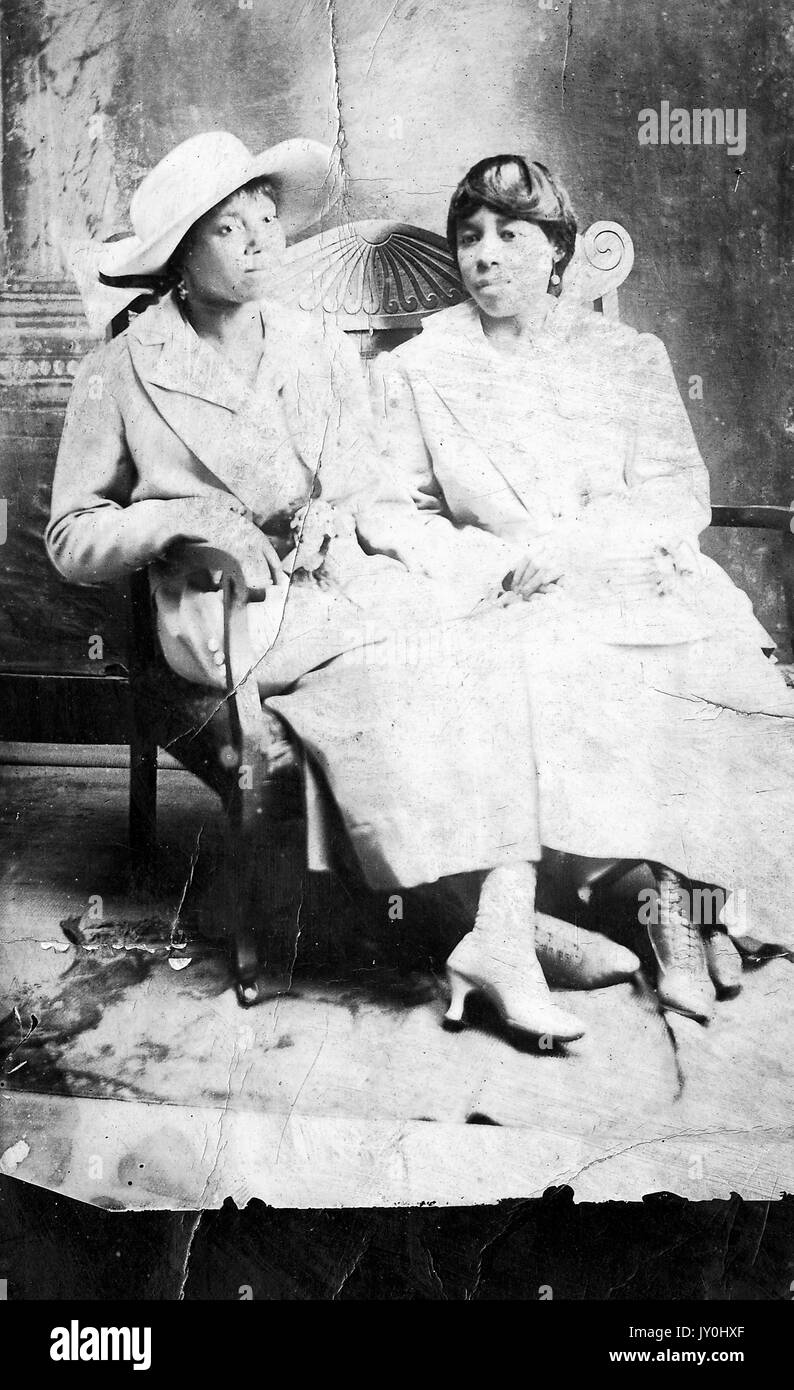 Sitzendes Portrait von zwei reifen afroamerikanischen Frauen, die leichte Kleider tragen, eine mit leichtem Hut, auf einer Bank vor der Kulisse sitzen, neutrale Ausdrücke, 1920. Stockfoto