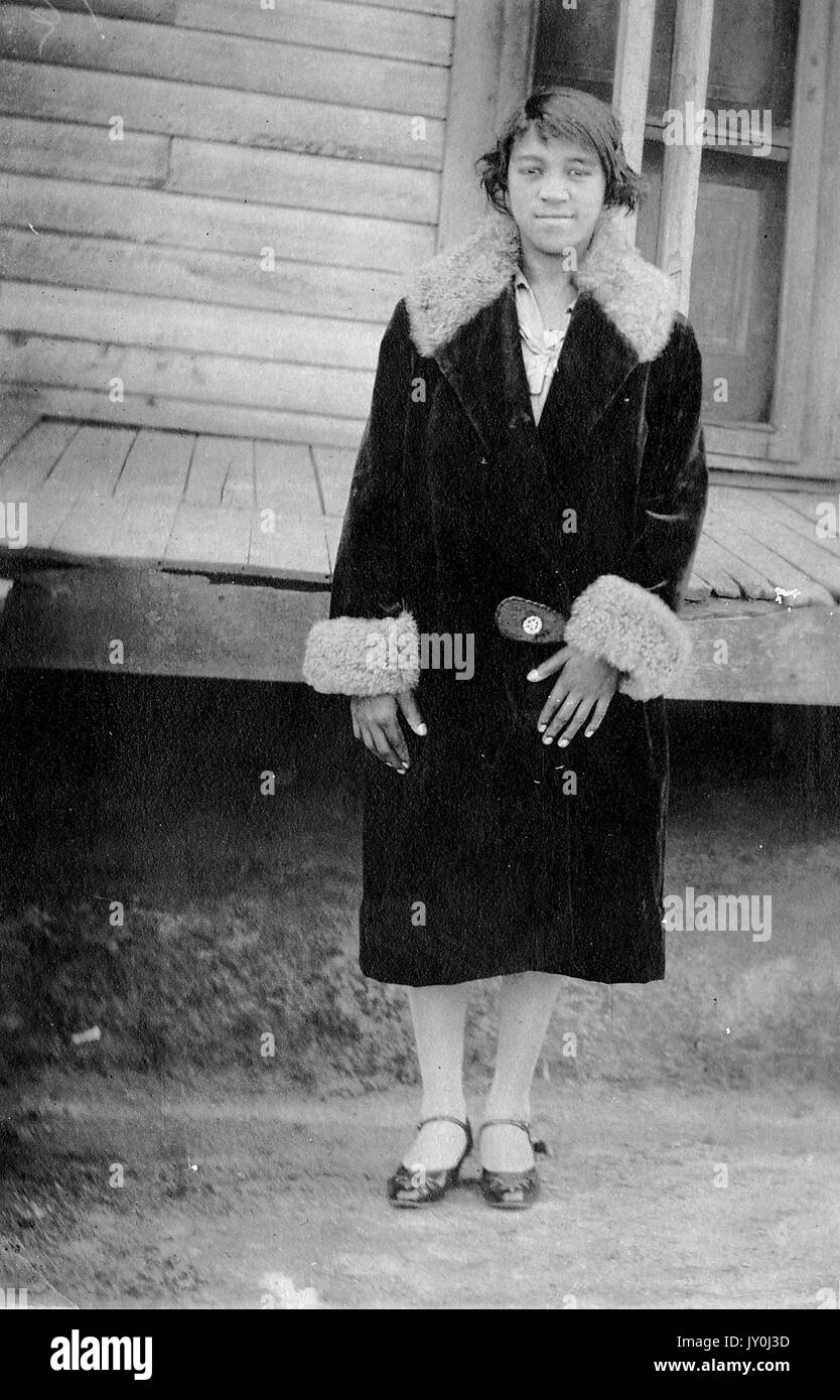 Porträt einer jungen afroamerikanischen Frau, sie steht vor einem Deck, sie ist sehr schön gekleidet, sie trägt einen langen dunklen Wintermantel, Strumpfhosen und schöne dunkelfarbige Kleid Schuhe, 1915. Stockfoto