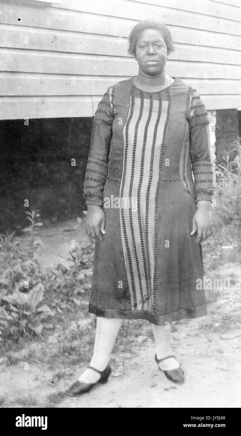 Porträt einer afroamerikanischen Frau, die vor einer erhöhten Struktur steht, sie ist schön gekleidet, trägt ein langes Kleid, Strumpfhosen und dunkelfarbige nahe gekleidete Schuhe, 1915. Stockfoto