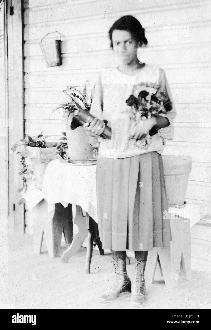 Porträt einer afroamerikanischen Frau namens Mrs ASP, sie steht auf einer Veranda und vor einem kleinen Tisch, der Blumentöpfe hält, sie hält ein Buch in einem Arm und Blumen in ihrem anderen Arm, Sie trägt ein lockeres, helles Hemd und einen langen, hellen Rock, sie trägt hohe Schnürstiefel, 1915. Stockfoto