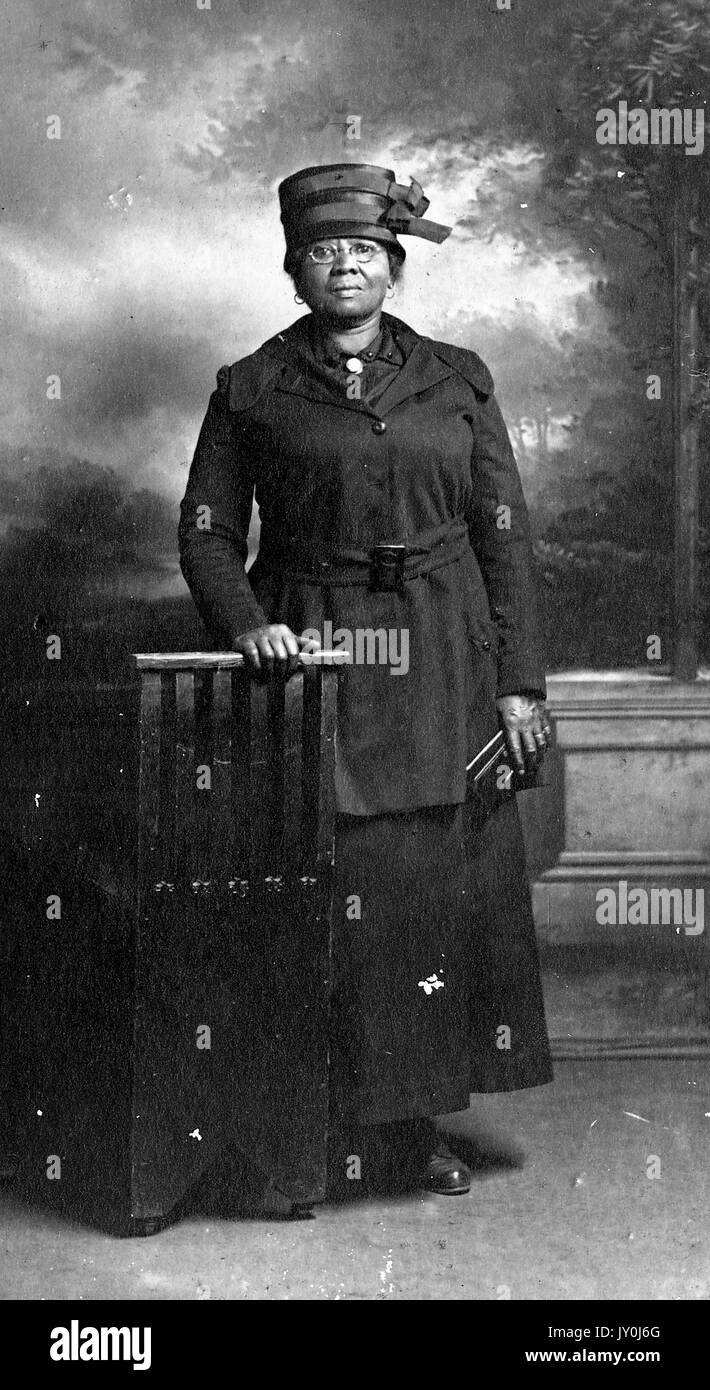 Portrait einer afrikanischen amerikanischen Frau namens Sallie Fitzjewel, sie trägt einen langen dunklen Trenchcoat und einen dunklen Hut, sie lehnt sich mit ihrer rechten Hand auf einer hölzernen Stütze und sie hält eine Kupplung in ihrer linken Hand, Nashville, Tenn, 1929. Stockfoto