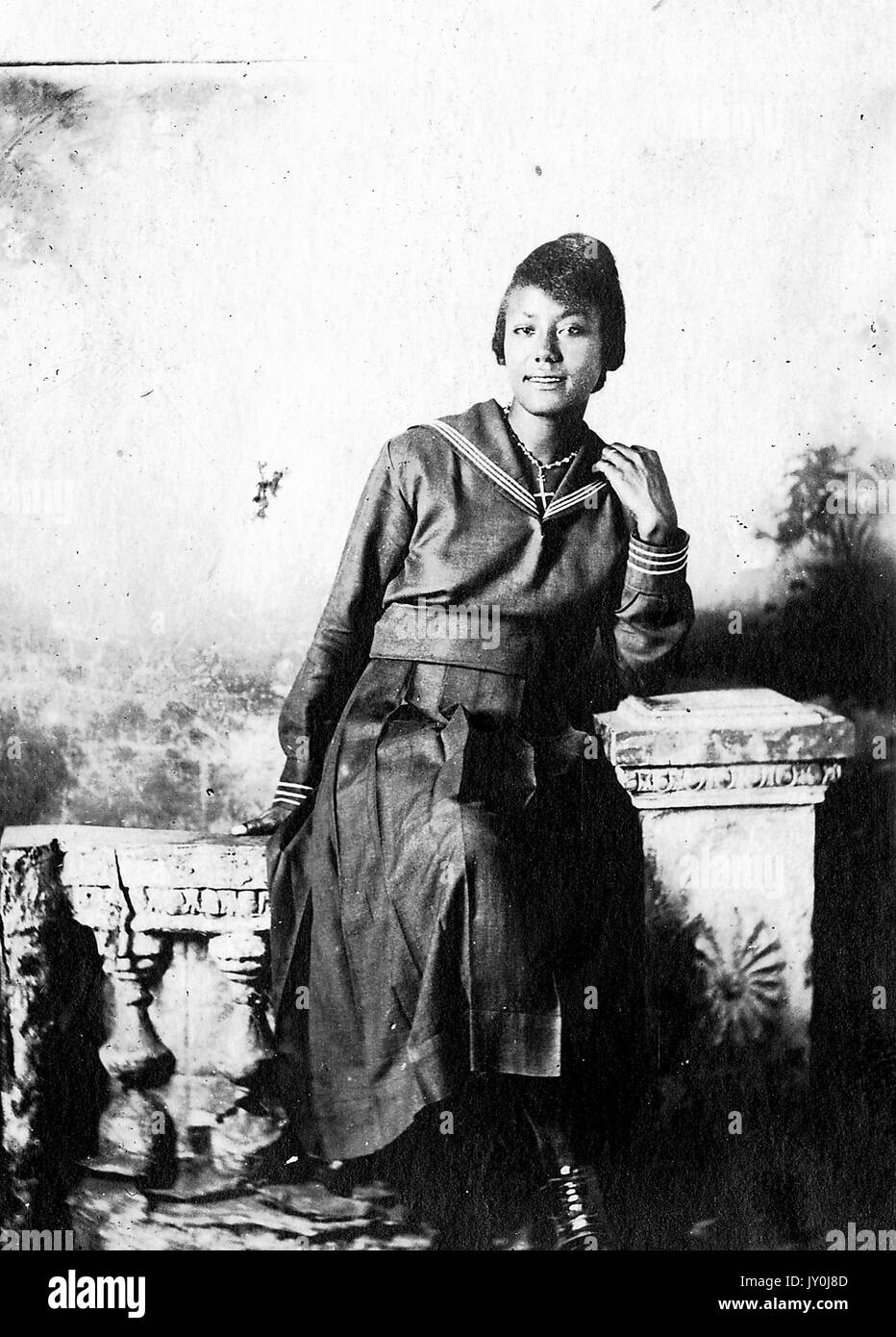 Porträt einer afroamerikanischen Frau, die auf Strukturrequisiten sitzt, einen langen dunklen Rock trägt und ein langärmeliges Oberteil trägt, ihren linken Ellbogen auf eine Struktursäule stützt, 1915. Stockfoto
