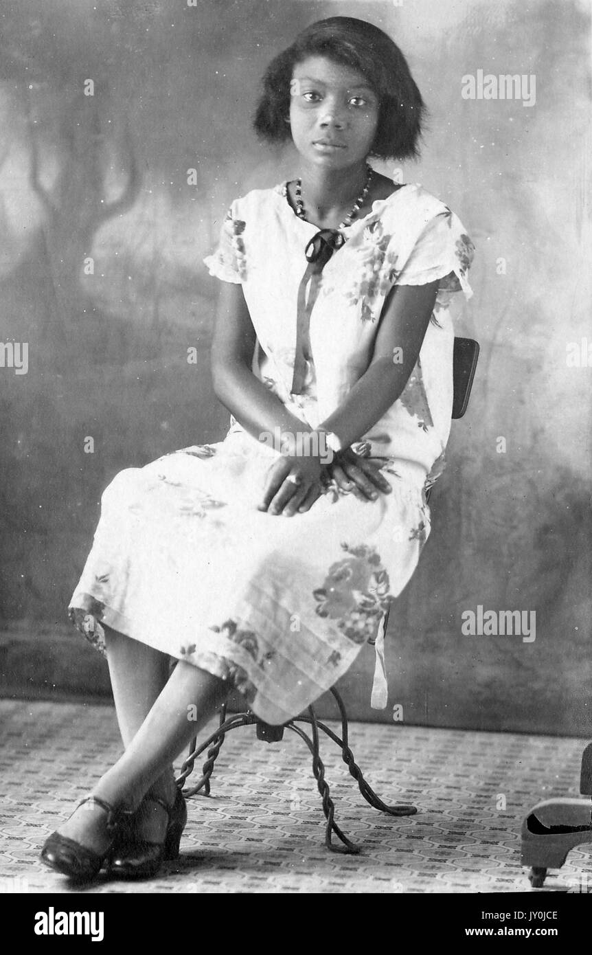 Porträt einer afroamerikanischen Frau, die auf einem Stuhl sitzt, sie trägt ein gemustertes Kleid und eine Halskette, sie ist in dunklen Kleidschuhen, sie trägt eine Uhr und einen Ring, ihre Hände sind gekreuzt und ruhen auf ihrem Schoß, 1915. Stockfoto