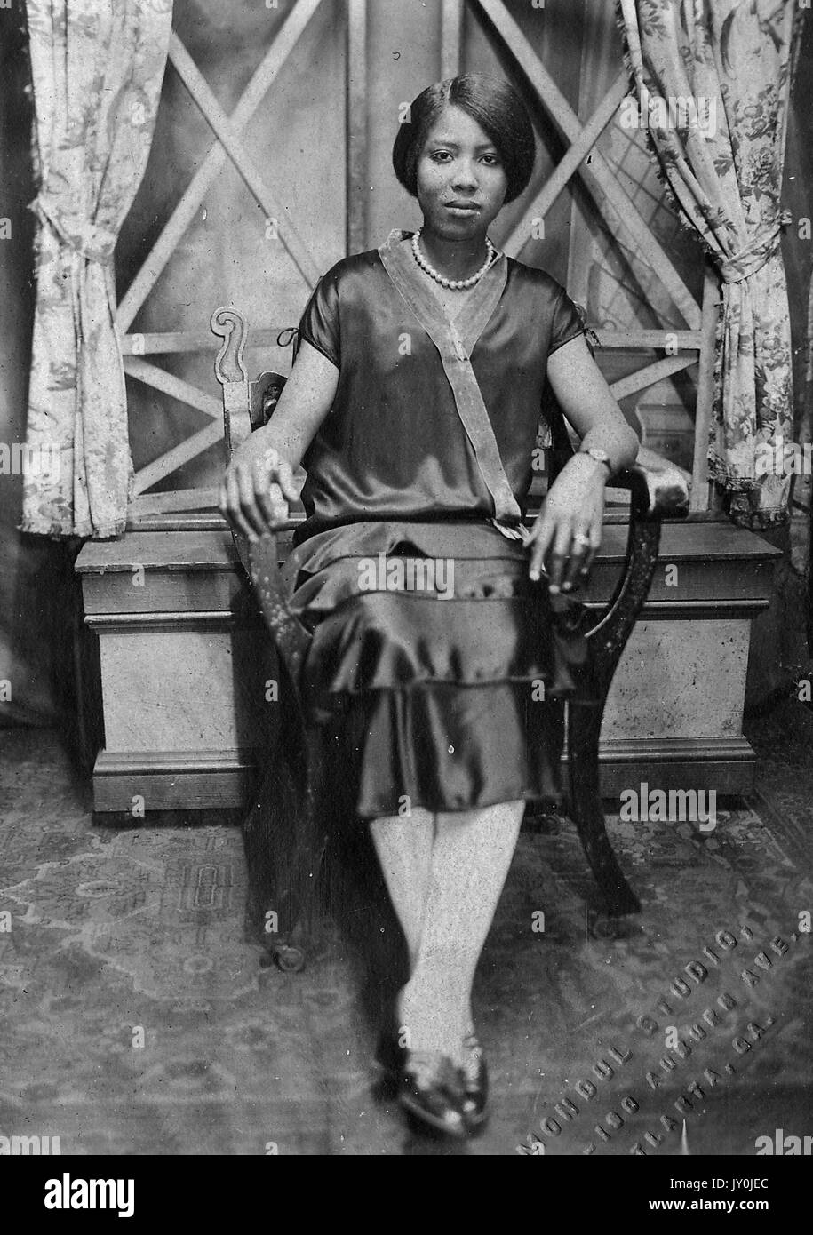 Porträt einer jungen afroamerikanischen Frau, die vor einem Fenster in einem Holzstuhl sitzt, ein dunkles Kleid und weiße Strumpfhosen trägt, ihre Arme auf den Armen des Stuhls ruhen, Atlanta, Georgia, 1915. Stockfoto