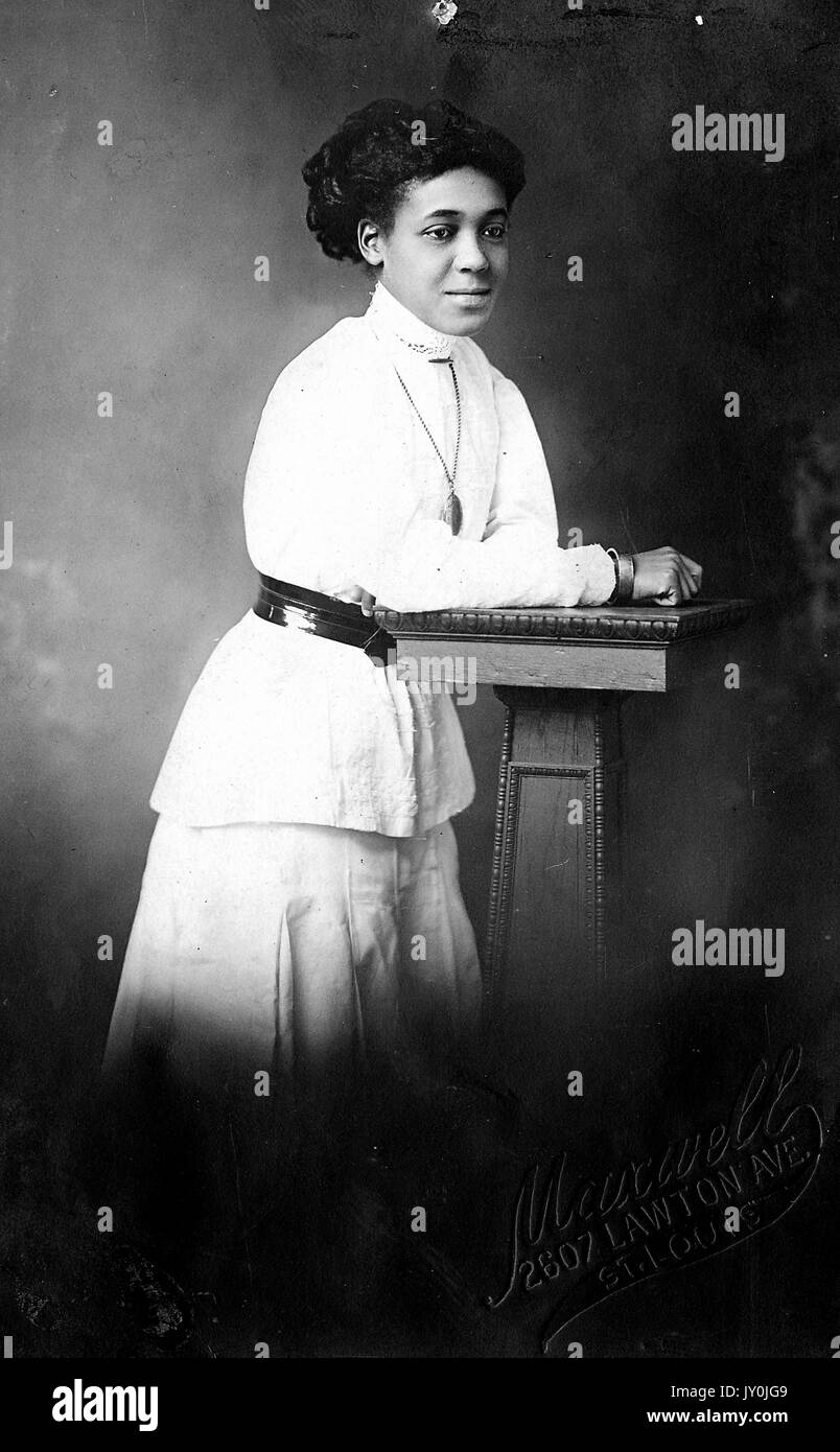 Porträt einer afroamerikanischen Frau, die auf einer Holzrequisite mit den Armen steht und einen weißen Rock und Hemd und einen dunklen Gürtel trägt, 2607 Lawton Ave, St. Louis, 1915. Stockfoto