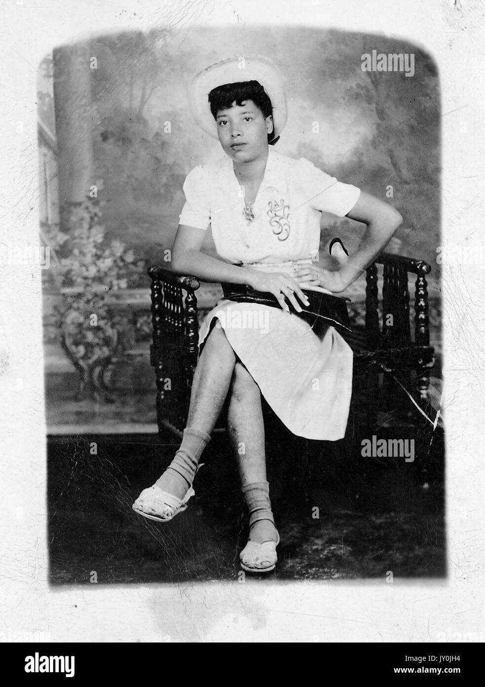 Porträt einer afroamerikanischen Frau, die mit gekreuzten Beinen auf einer Bank sitzt, ein helles Kleid und einen Hut trägt, ihre rechte Hand hält eine dunkle Geldbörse in ihrem Schoß, ihr linker Arm ist auf ihrer Hüfte, 1915. Stockfoto