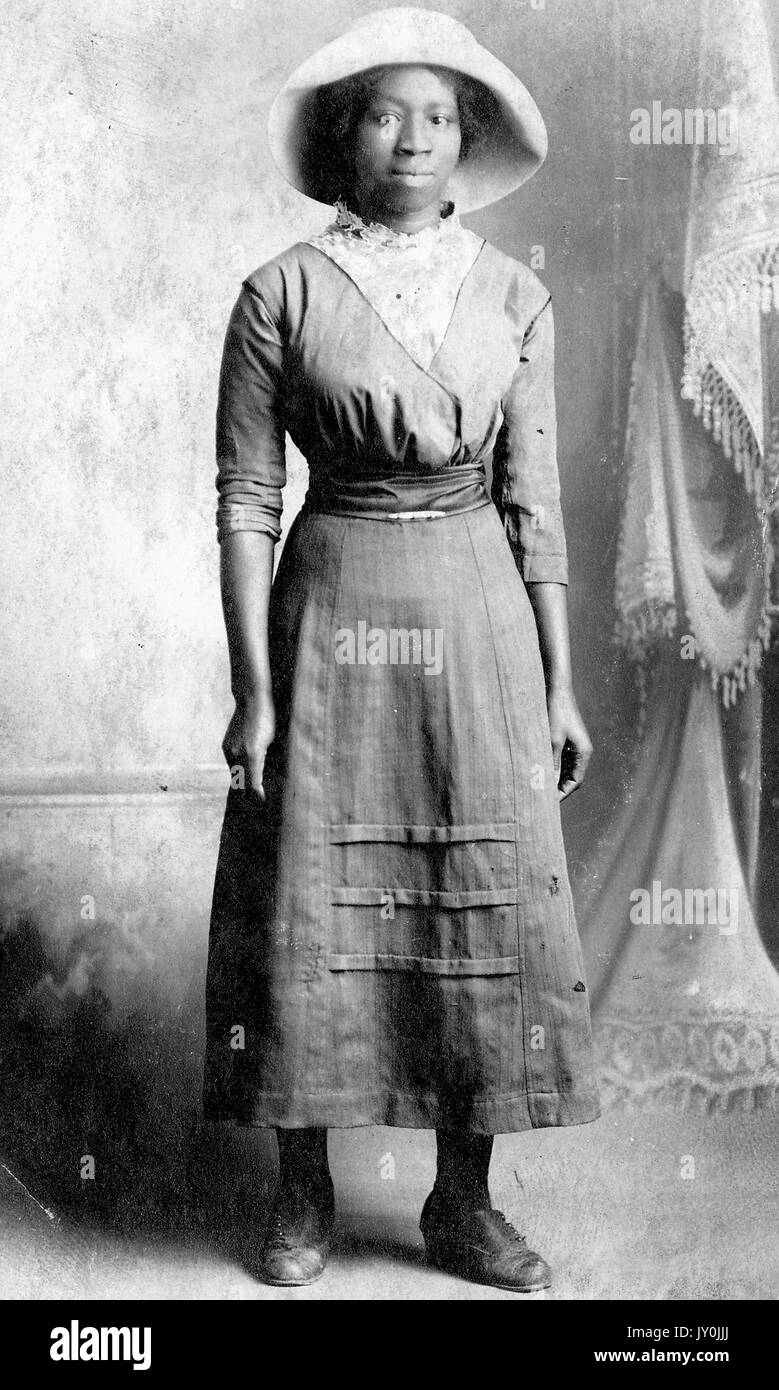 Portrait einer afrikanischen amerikanischen Frau, die vor einem Vorhang, Sie trägt ein langes Kleid, das in der Taille gegürtet ist, sie trägt einen hellen Hut und ihre Hände sind durch ihre Seiten, 1915. Stockfoto