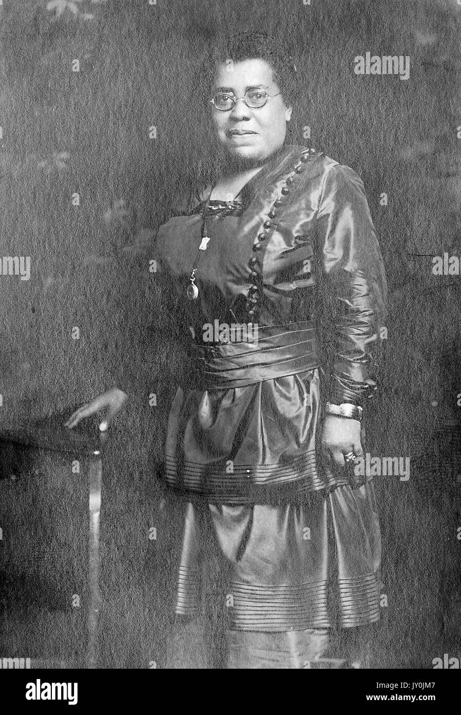 Porträt einer afroamerikanischen Frau, die mit ihrem rechten Arm eine Holzrequisite berührt, ein dunkles langärmeliges Kleid und eine Brille trägt, ihr linker Arm neben ihr, Sissan Studio, 1828 Center Ave, Pittsburg, PA, 1915. Stockfoto