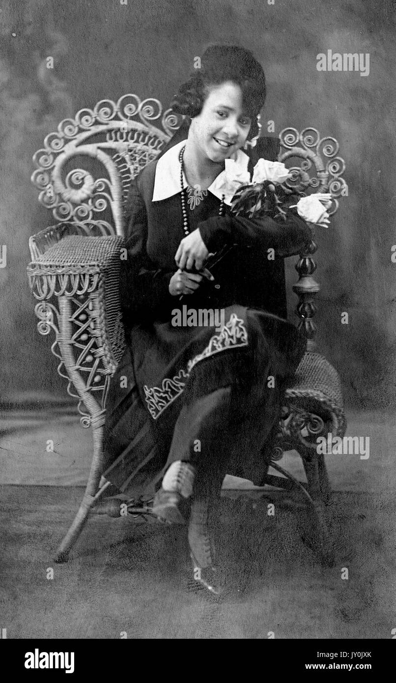 Porträt einer jungen afroamerikanischen Frau, die in einem Korbsessel sitzt, ihre Beine sind gekreuzt und sie hält einen Blumenstrauß in ihrem linken Arm, sie trägt ein dunkles Outfit mit einem großen hellen Kragen, sie lächelt, 1915. Stockfoto