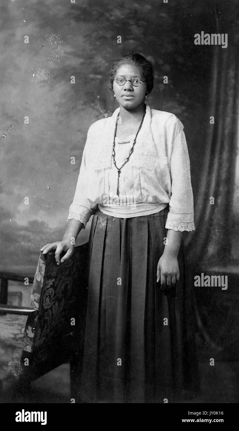 Porträt einer afroamerikanischen Frau, die vor einem Wandbild und einem Vorhang steht, sie trägt einen dunklen langen Rock und eine helle Bluse, sie trägt eine Halskette, eine Brille, Und Ohrringe, ihr rechter Arm ruht auf einem Stuhl und ihr linker Arm ist neben ihr, 1915. Stockfoto