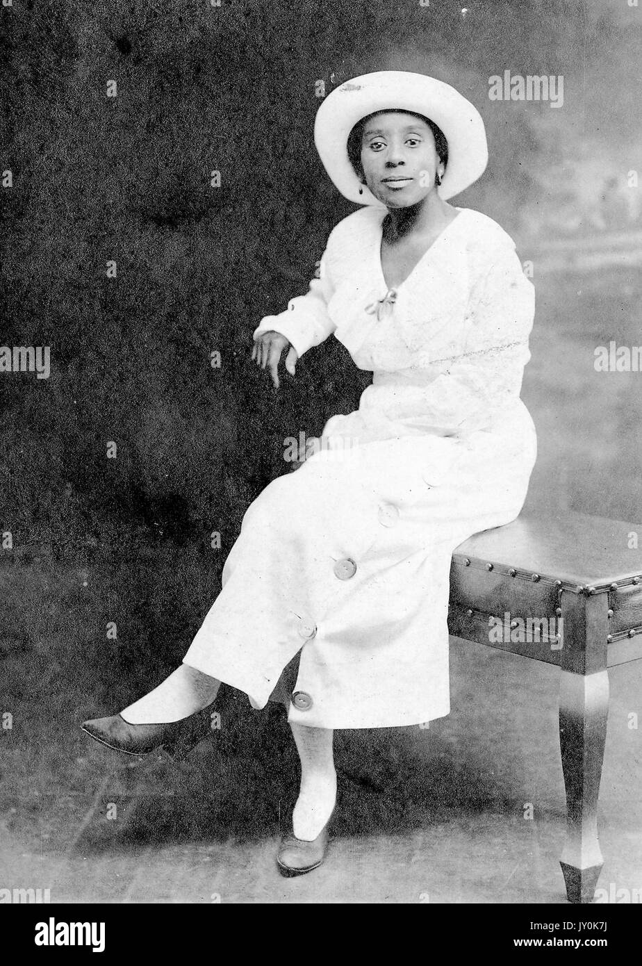 Sitzendes Porträt einer reifen afroamerikanischen Frau in hellem Kleid, Ohrringen und Hut, auf Bank vor Hintergrund sitzend, neutraler Ausdruck, 1920. Stockfoto