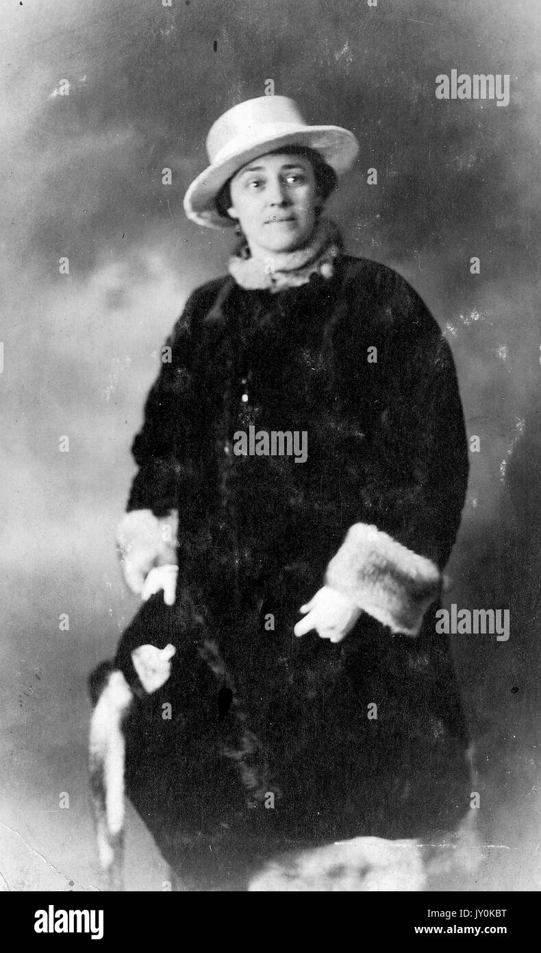 Drei Viertel Länge stehend Porträt der reifen afroamerikanischen Frau, trägt dunklen Mantel mit Pelzdetails, Hut und aufwendigen Kragen, Geldbörse, neutraler Ausdruck, 1920. Stockfoto