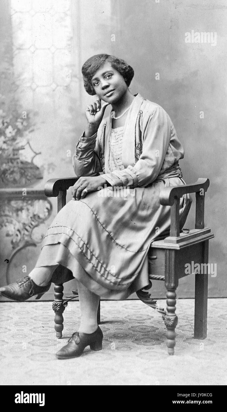 Ganzkörperportrait der jungen afroamerikanischen Frau, in leichtem Kleid, Halskette und Ringen, auf einem kunstvollen Holzstuhl vor der Kulisse sitzend, neutraler Ausdruck, 1925. Stockfoto