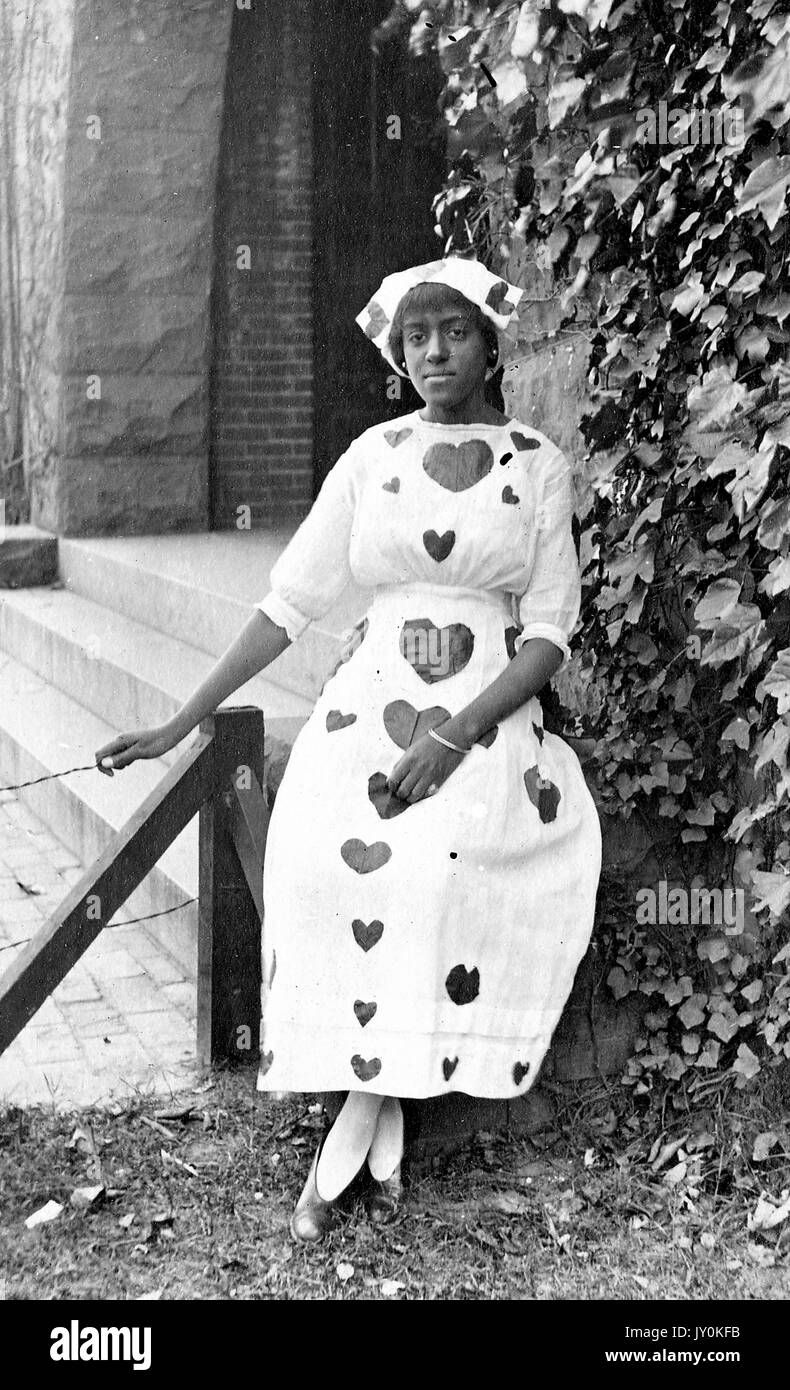 Sitzendes Portrait einer jungen afroamerikanischen Frau, in hellem Kleid mit dunklen Herzen und Mütze mit passenden Designs, außerhalb des Gebäudes neben Pflanzen stehend, neutraler Ausdruck, 1915. Stockfoto