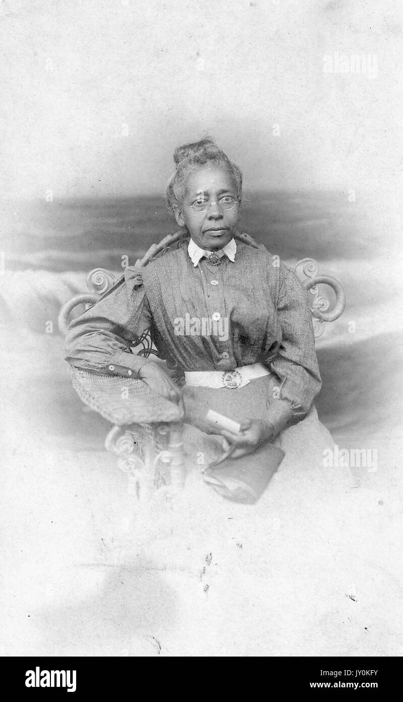 Dreiviertel sitzendes Porträt einer älteren afroamerikanischen Frau, die dunkles Kleid mit hellem Kragen, Gürtel und Brille trägt, Geldbörse und Papier hält, neutraler Ausdruck, 1915. Stockfoto