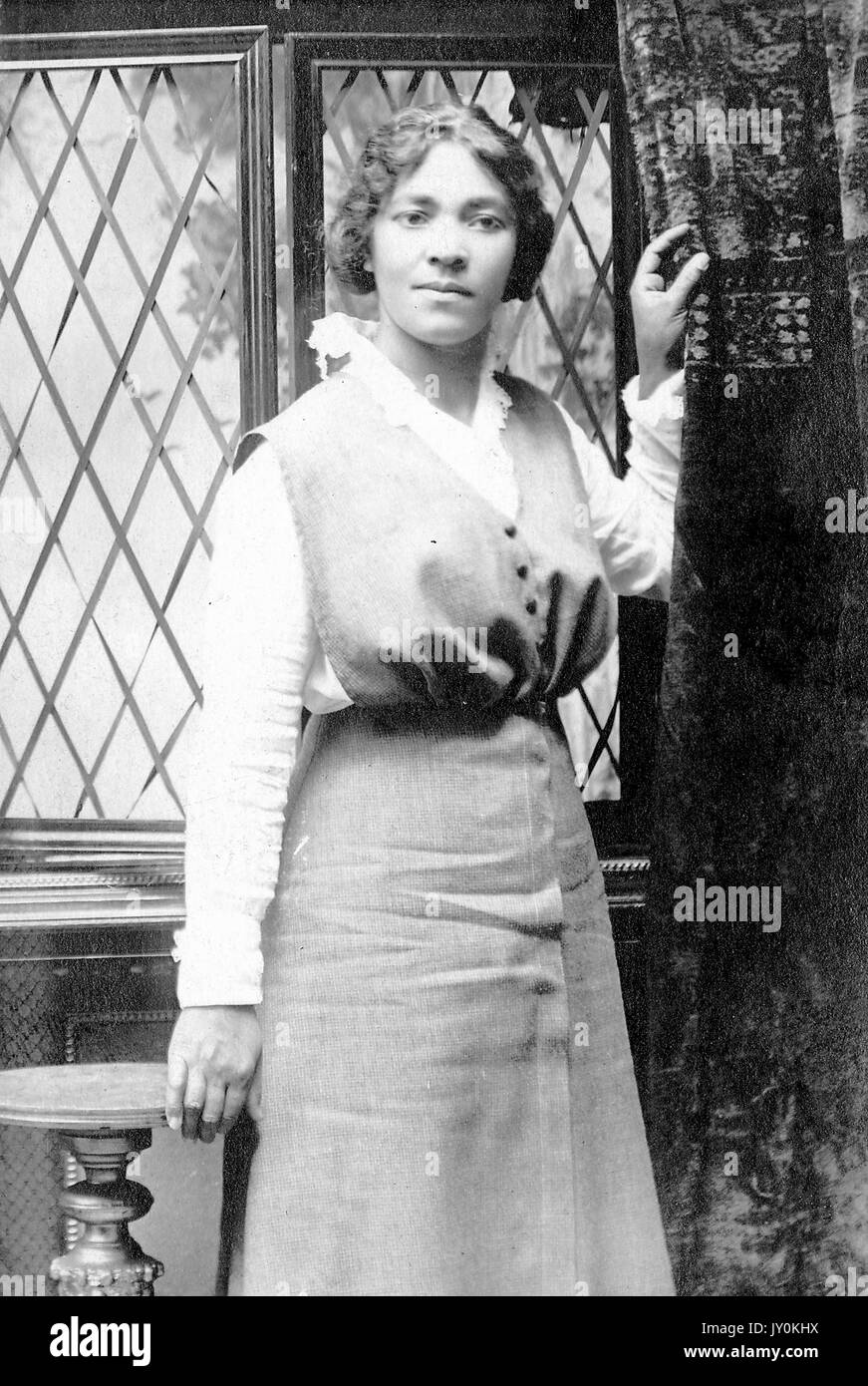 Drei Viertel lang stehend Porträt der jungen afroamerikanischen Frau, tragen dunkle Kleid über helle Bluse, vor dem Fenster neben stehen, während Vorhang halten, neutral Ausdruck, 1920. Stockfoto