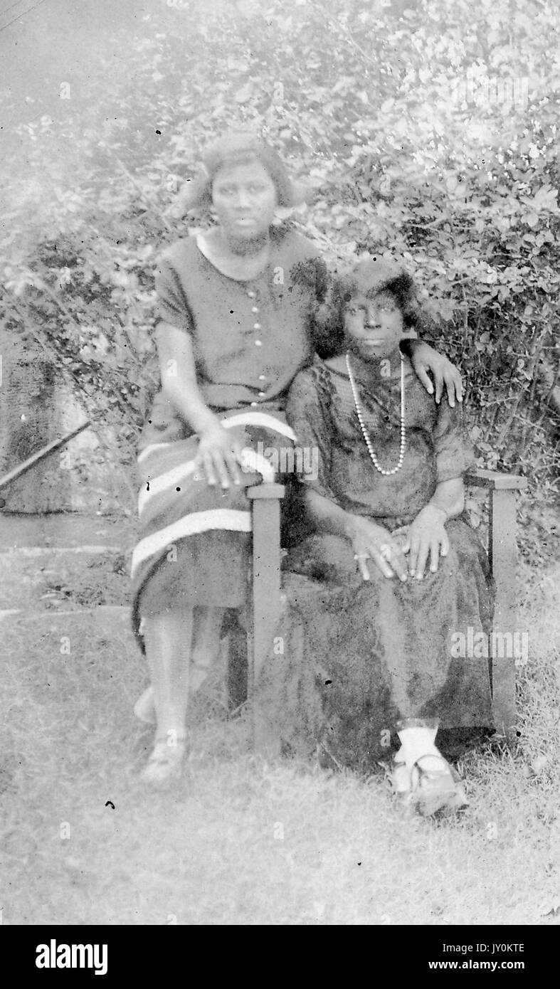 Sitzendes Porträt zweier afroamerikanischer Frauen in voller Länge, eine Frau trägt dunkles Kleid mit hellem Detail, eine andere Frau trägt dunkles Kleid mit Halskette, draußen sitzend mit Gras, 1920. Stockfoto