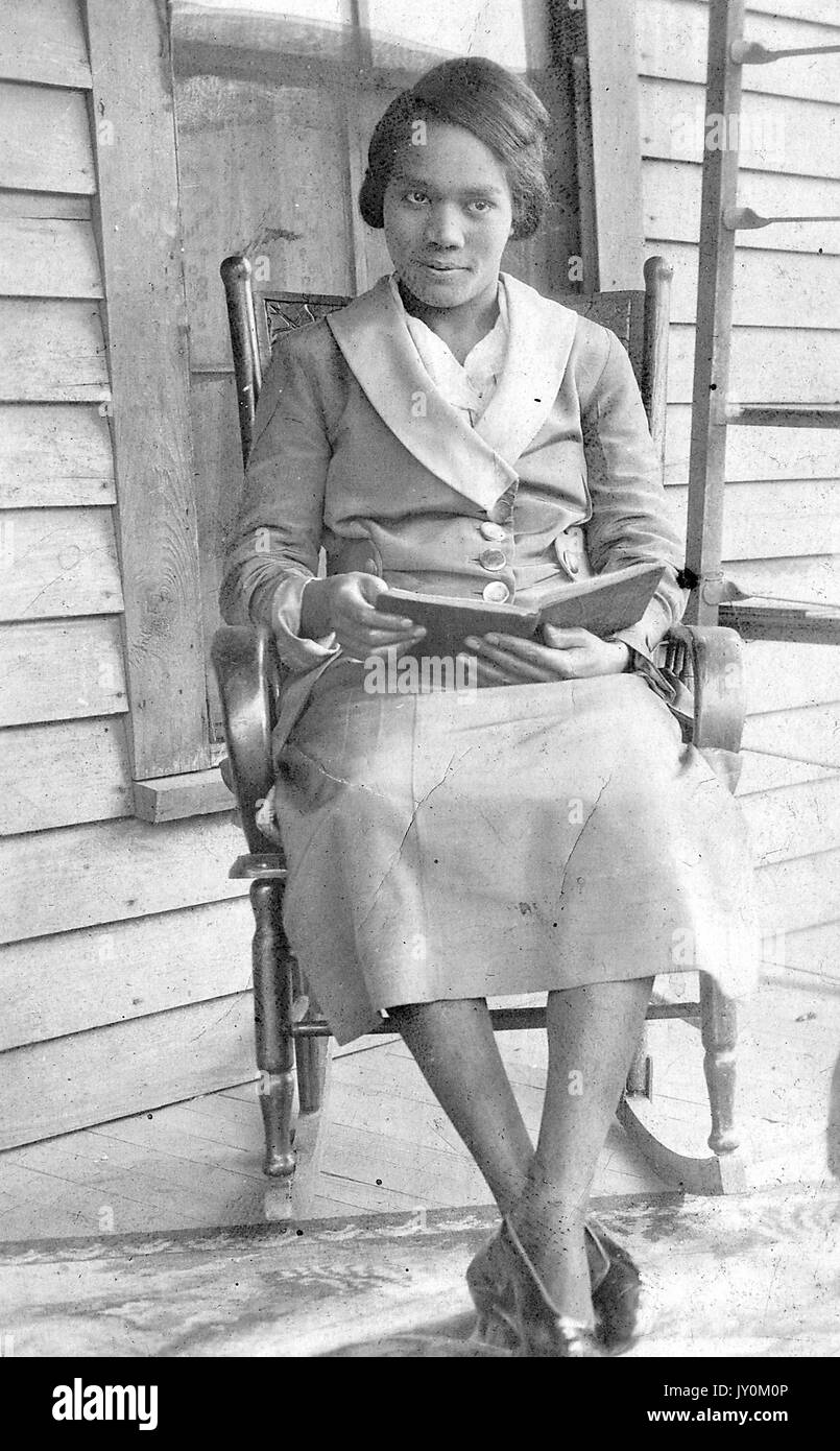 Sitzendes Portrait der reifen afroamerikanischen Frau, in leichtem Kleid mit Blazer, offenes Buch haltend, neutraler Ausdruck, auf Veranda sitzend auf Schaukelstuhl, 1915. Stockfoto