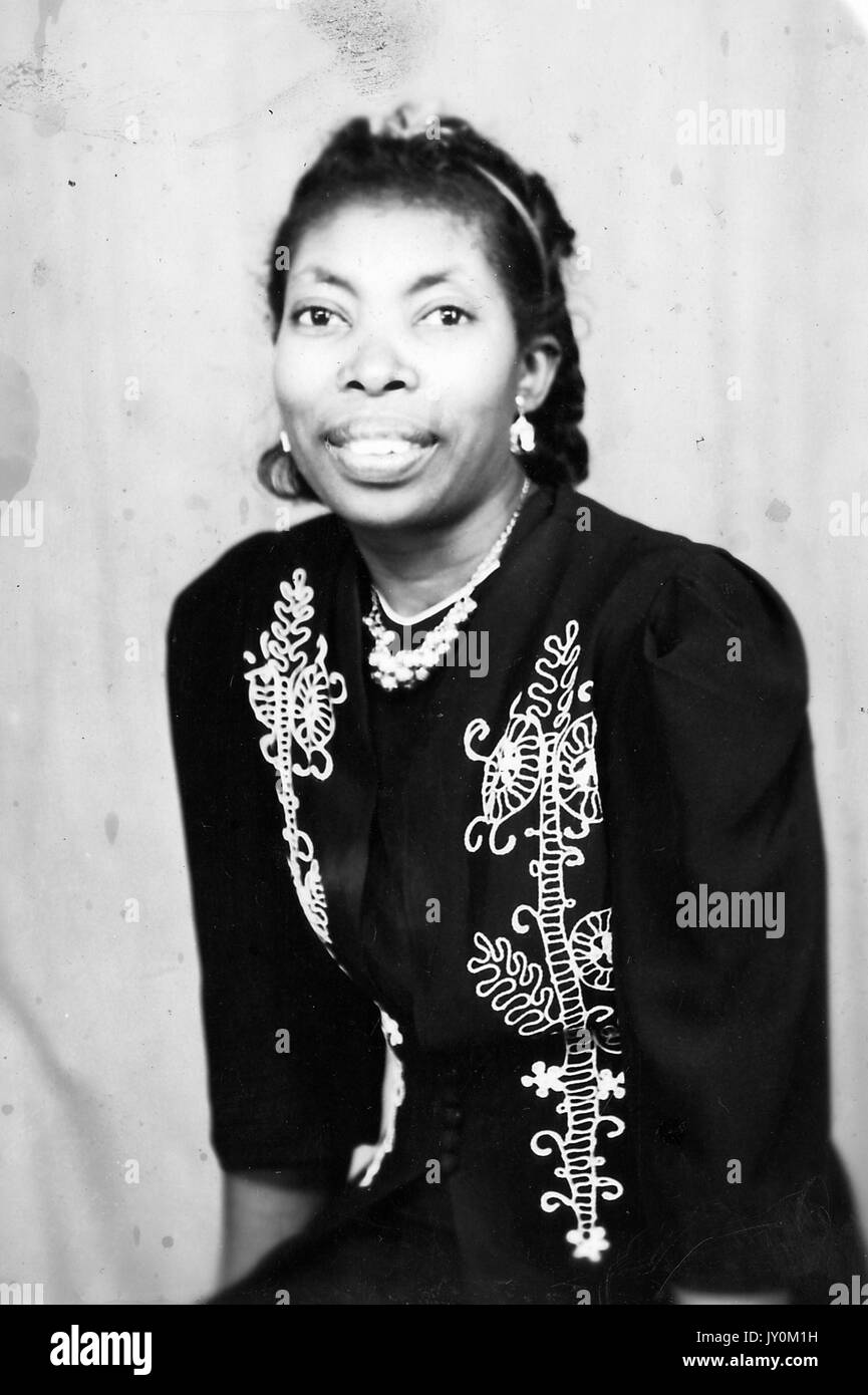 Halblanges sitzendes Porträt einer reifen afroamerikanischen Frau, in dunklem Kleid mit floralen Details, Halskette, Ohrringen und Stirnband, lächelnder Ausdruck, 1920. Stockfoto