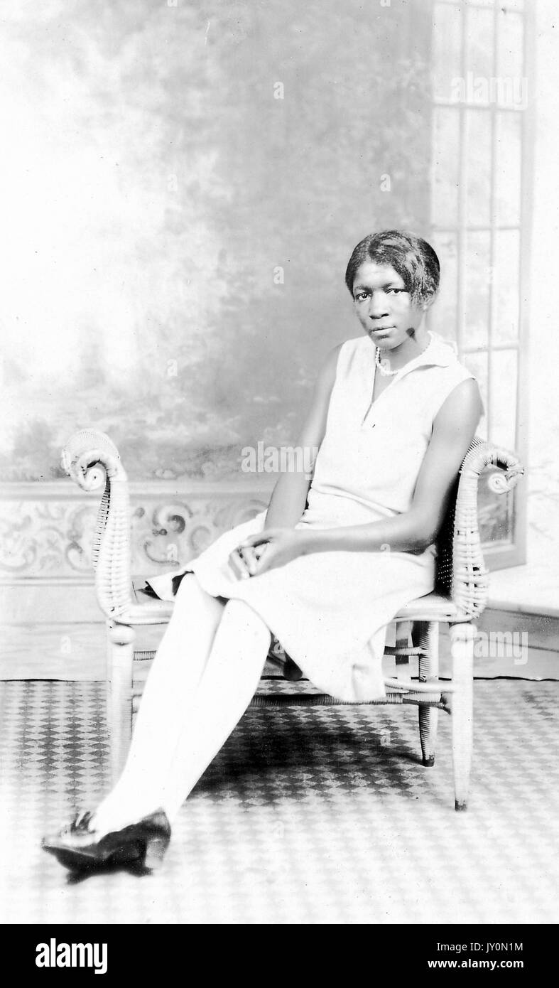 Ganzkörperportrait einer sitzenden afroamerikanischen Frau, die ein weißes Kleid und weiße Strümpfe trägt, die Hände auf dem Schoß gefaltet, eine Perlenkette trägt, auf einem Holzstuhl vor der Kulisse sitzt, 1920. Stockfoto
