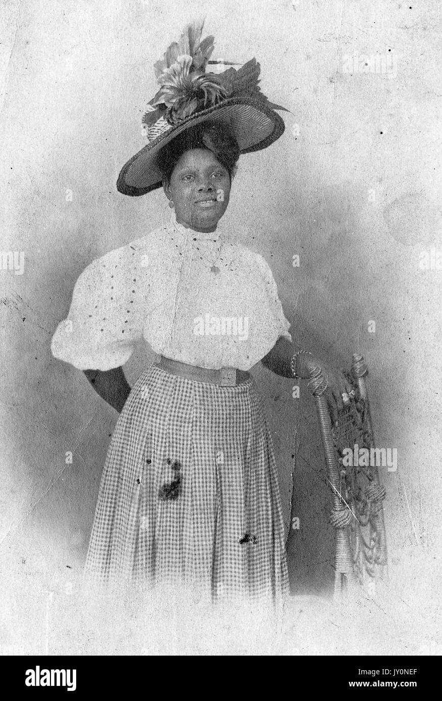 Dreiviertel-Porträt einer afroamerikanischen Frau, die neben einem Stuhl steht, mit einer Hand auf dem Stuhl, mit einem karierten Kleid und einer weißen Bluse, mit einem verzierten Federhut auf dem Kopf, mit einem lächelnden Gesichtsausdruck, 1920. Stockfoto