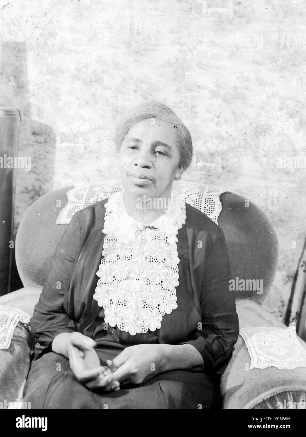 Halbkörperportrait einer afroamerikanischen Frau, die auf einem Armstuhl sitzt, die Hände auf dem Schoß trägt, ein dunkles Kleid trägt, vor einer Kulisse, mit einem ernsten Gesichtsausdruck, 1920. Stockfoto