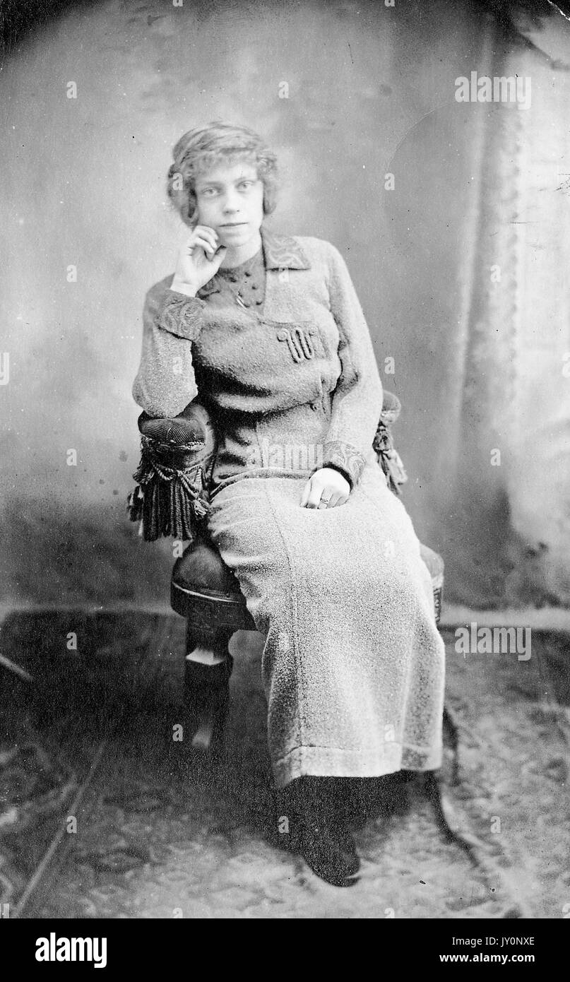 Ganzkörperportrait einer afroamerikanischen Frau, die auf einem Stuhl vor einer Kulisse saß, eine Hand auf ihrem Schoß, die andere Hand auf ihrem Gesicht ruhte, ein dunkles Kleid trug, mit einem ernsten Gesichtsausdruck, 1920. Stockfoto