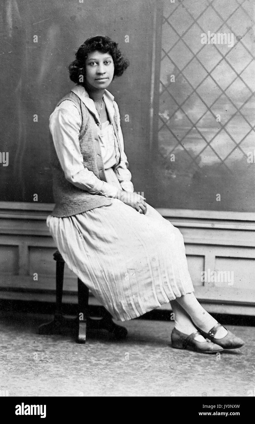 Ganzkörperportrait, junge afroamerikanische Frau, in leichtem Kleid und Weste, auf Hocker vor Hintergrund sitzend, neutraler Ausdruck, 1920. Stockfoto