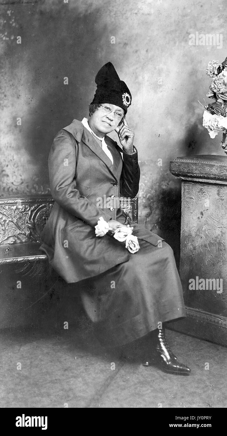 Sitzendes Porträt einer reifen afroamerikanischen Frau auf einer Bank, in dunklem Kleid, dunklem Blazer und Hut mit Verzierungen, mit hellen Blumen, neutraler Ausdruck, 1915. Stockfoto