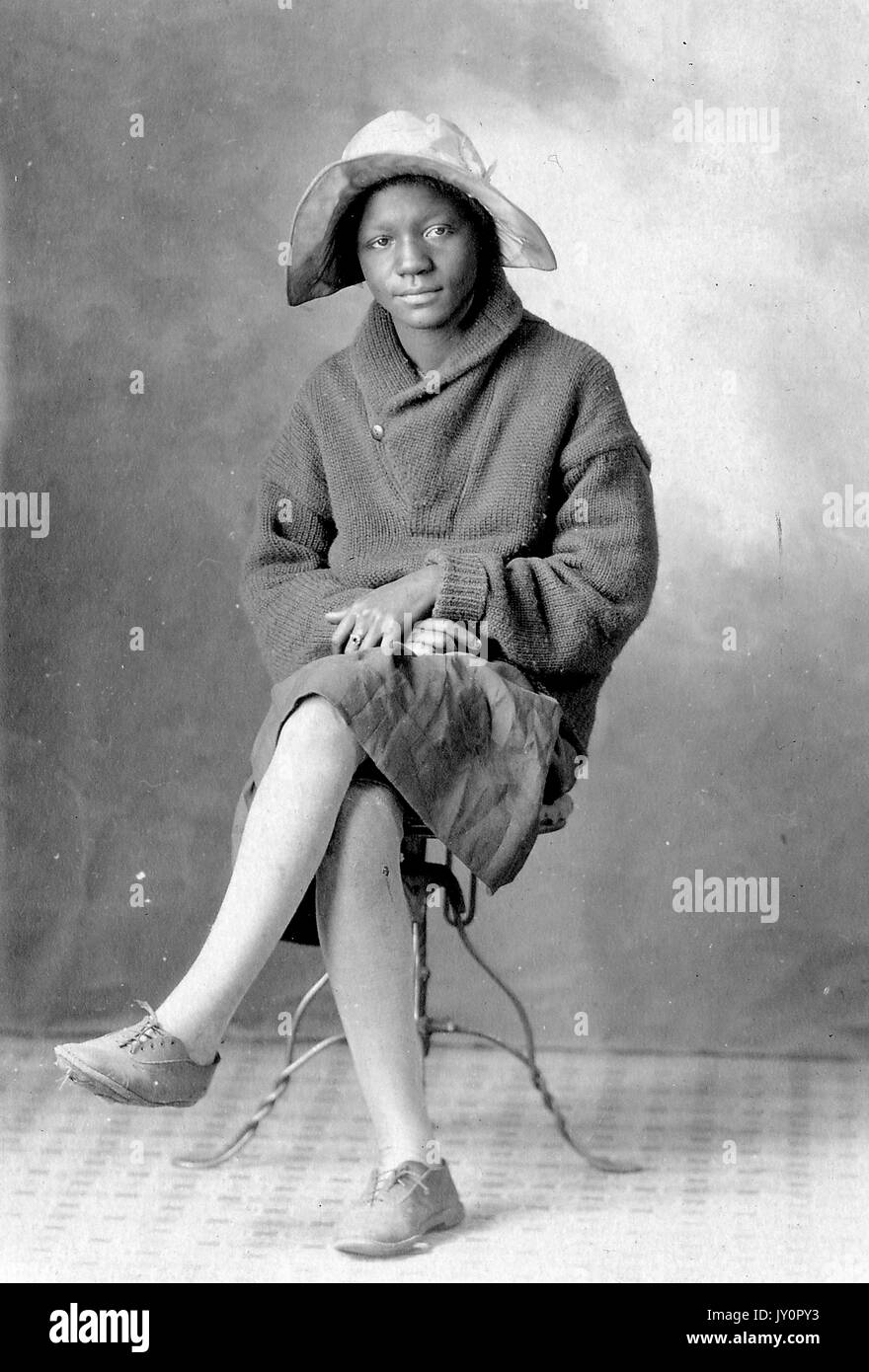 Volle Länge sitzen Porträt, junge afrikanische amerikanische Frau sitzt auf einem Stuhl, tragen dunkle Kleidung, dunklen pullover und Hut, neutralen Ausdruck, 1925. Stockfoto