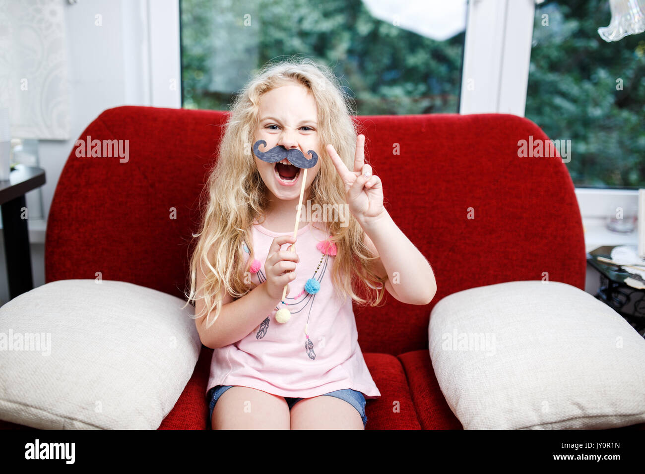 Süße kleine Mädchen mit Papier Schnurrbärte sitzend auf roten Stuhl zu Hause. Stockfoto