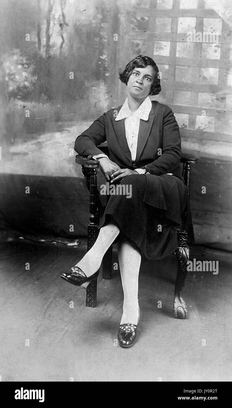 Volle Länge sitzen Porträt, junge afrikanische amerikanische Frau, tragen dunkle Kleidung mit hellen Bluse, sitzen vor dem Hintergrund, neutralen Ausdruck, 1920. Stockfoto