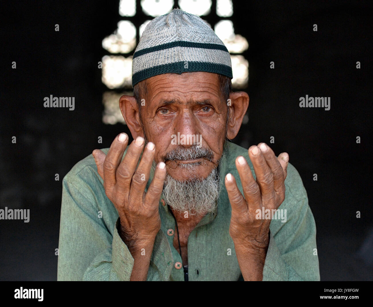 Der alte indische Moslem-Mann mit gelebtem Gesicht trägt eine islamische Betkappe (taqiyah) und betet mit beiden Händen. Stockfoto