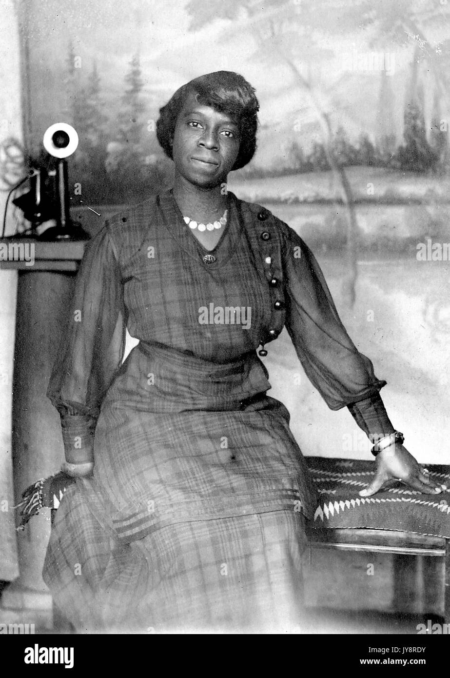 Dreiviertel sitzendes Porträt einer reifen afroamerikanischen Frau, die auf einer zerbrochenen Bank vor einer bemalten Kulisse und neben einem Telefon sitzt, ein kariertes Kleid, eine Halskette, ein Armband und einen neutralen Ausdruck trägt, 1915. Stockfoto