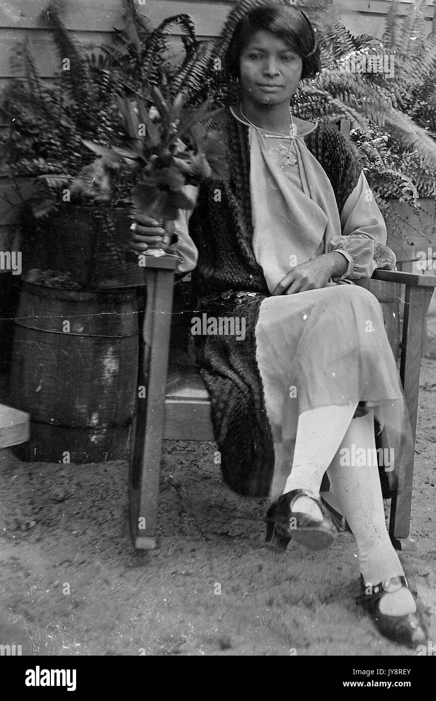 Volle Länge sitzt Porträt der jungen afrikanischen amerikanischen Frau, draußen sitzen vor einem Gebäude und Pflanzen, Pflanzen hält in der rechten Hand trägt ein Kleid, eine Halskette, Ohrringe, und dunkle Schuhe, 1915. Stockfoto