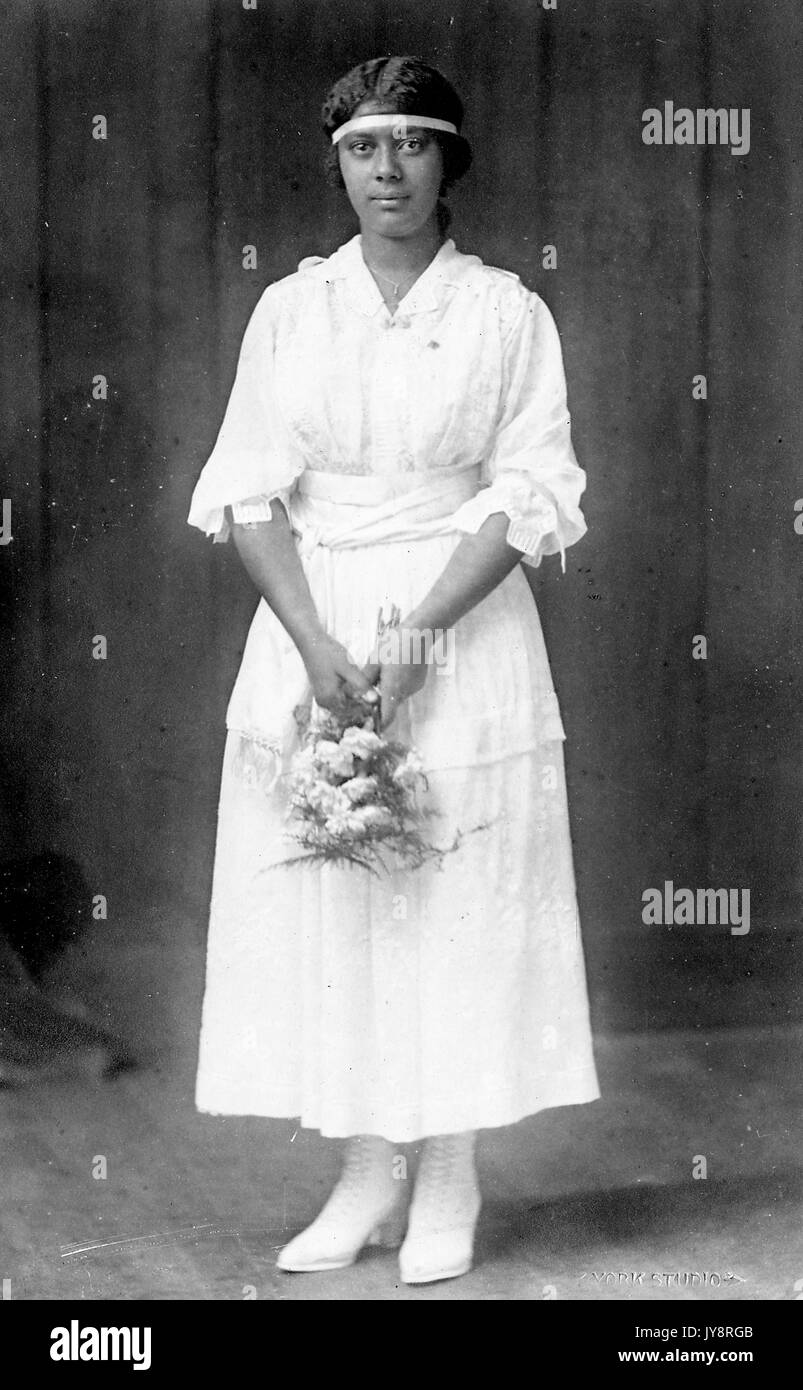 Volle Länge ständigen Porträt der jungen Afrikanische amerikanische Frau, die einen Blumenstrauß, trägt ein leichtes Kleid, Kopfband und einem neutralen Ausdruck, 1915. Stockfoto