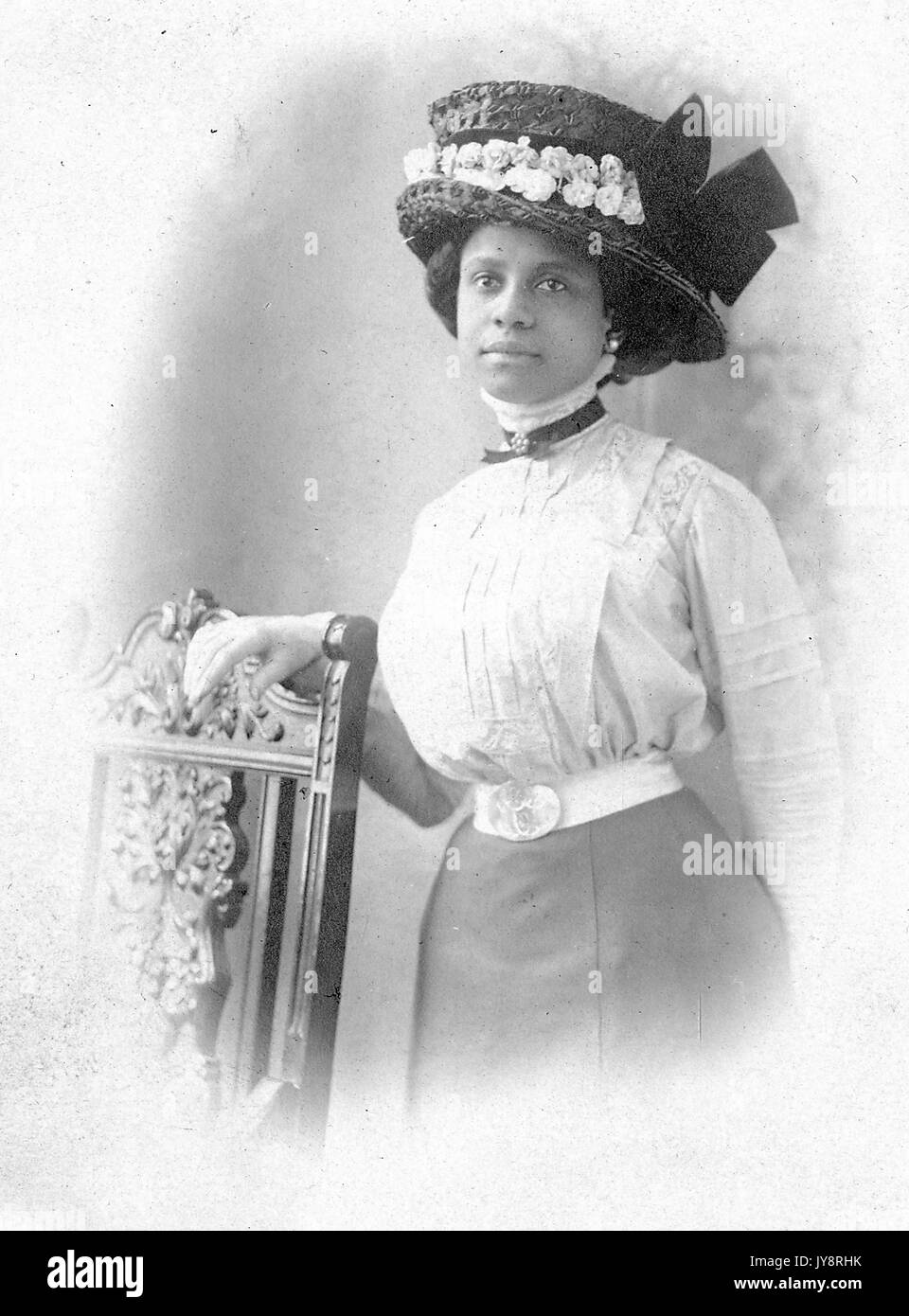 Halblanges Standbild einer jungen afroamerikanischen Frau neben dem Stuhl, die eine Bluse, einen Rock, einen aufwendigen Hut, Ohrringe und einen neutralen Ausdruck trägt, möglicherweise Philadelphia, Pennsylvania, 1915. Stockfoto