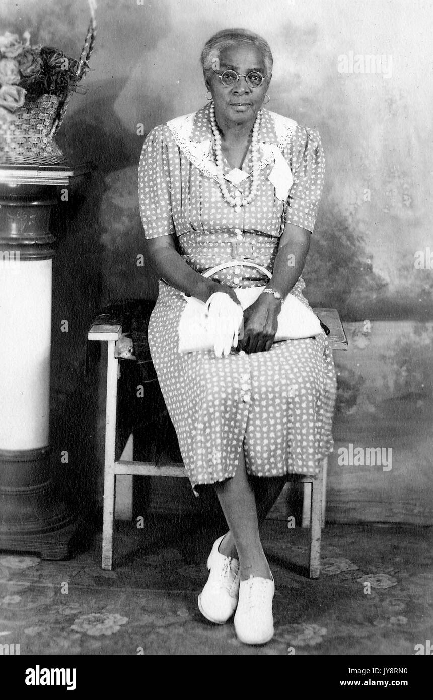 Volle Länge sitzt Portrait von älteren Afrikanische amerikanische Frau, sitzt vor einem gemalten Hintergrund neben einer Säule mit einem Korb mit Blumen, trägt ein gemustertes Kleid, Gläser, eine Halskette, Ohrringe, und Schnürung Schuhe, eine Handtasche, 1915. Stockfoto