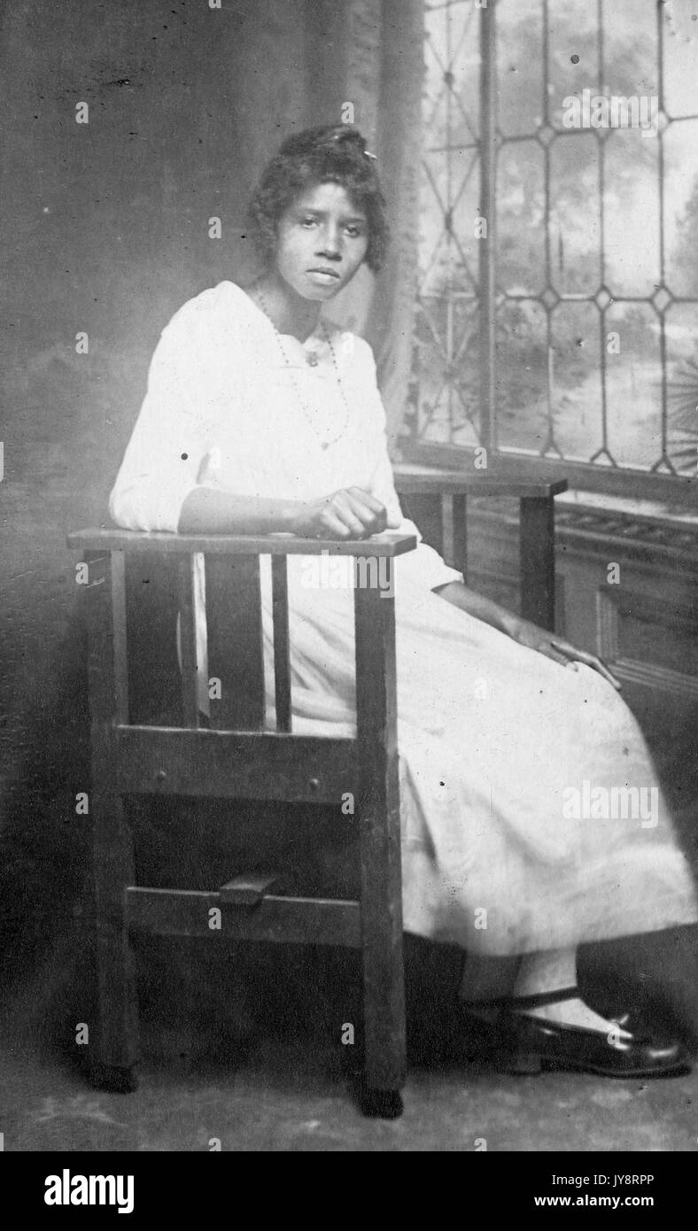 Volle Länge sitzt Porträt der afrikanischen amerikanischen Frau vor einem Fenster, trägt ein leichtes Kleid, eine Halskette und ein ernster Ausdruck, Nashville, Tennessee, 1915. Stockfoto