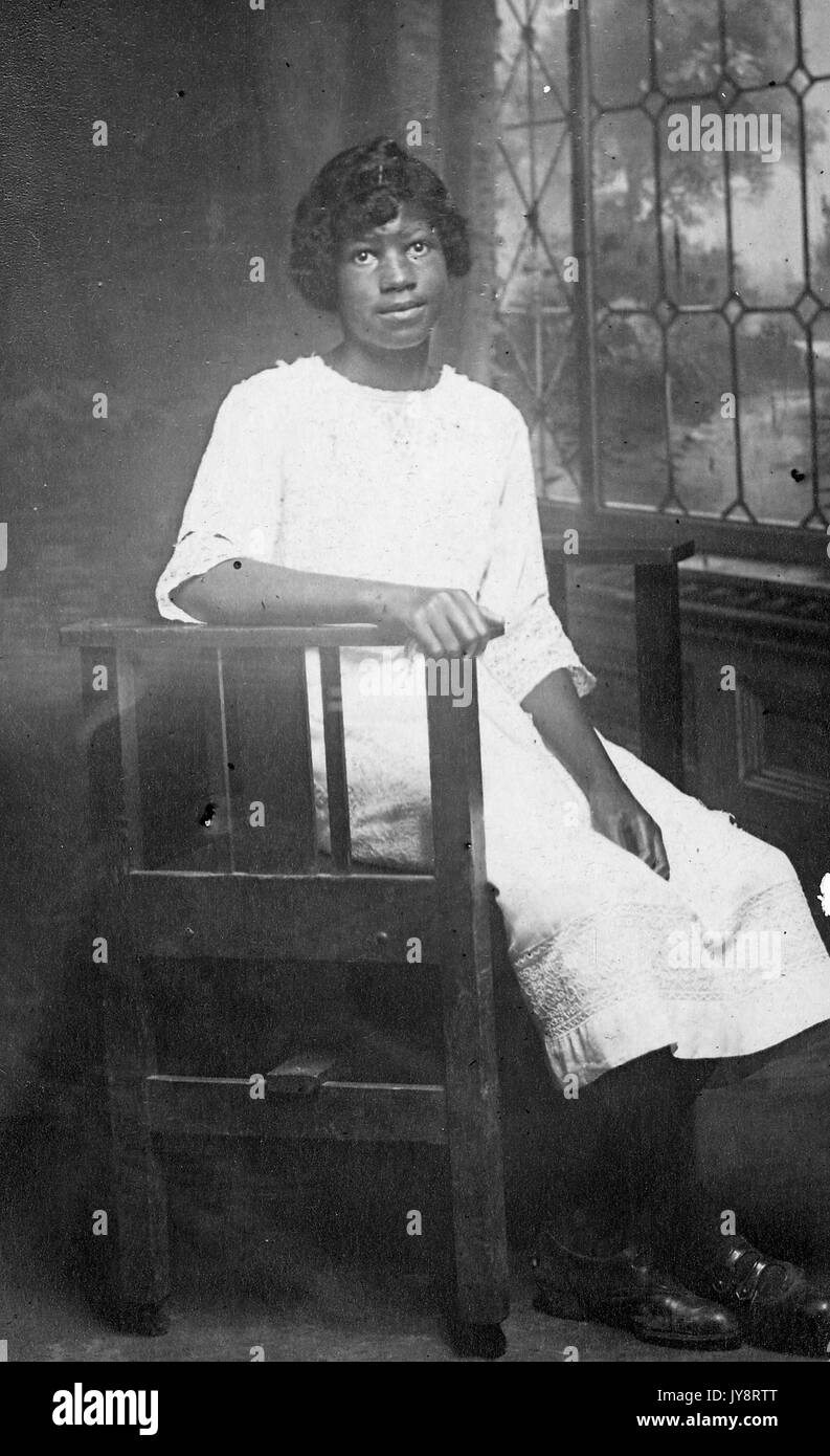 Volle Länge sitzt Porträt der jungen afrikanischen amerikanischen Frau vor einem Fenster, trägt ein leichtes Kleid und neutralen Ausdruck, möglicherweise in Nashville, Tennessee, 1915. Stockfoto