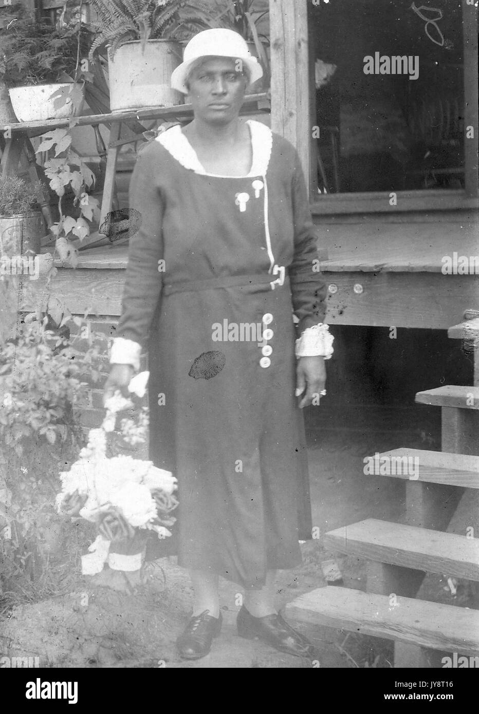 Ganzkörperportrait einer reifen afroamerikanischen Frau, die in der Nähe von Holztreppen steht, die zur Veranda eines Hauses führen, einen Blumenstrauß mit einem ernsten Gesichtsausdruck hält, 1915. Stockfoto