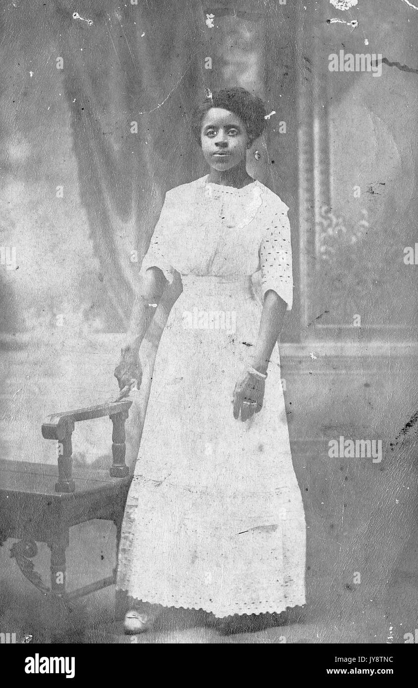 In voller Länge Porträt der jungen Afrikanische amerikanische Frau, die als Miss Redd, trug ein weißes Kleid und stand vor einer gemalten Kulisse, mit einem Finger auf den Arm einer Bank, 1944 ausruhen. Stockfoto