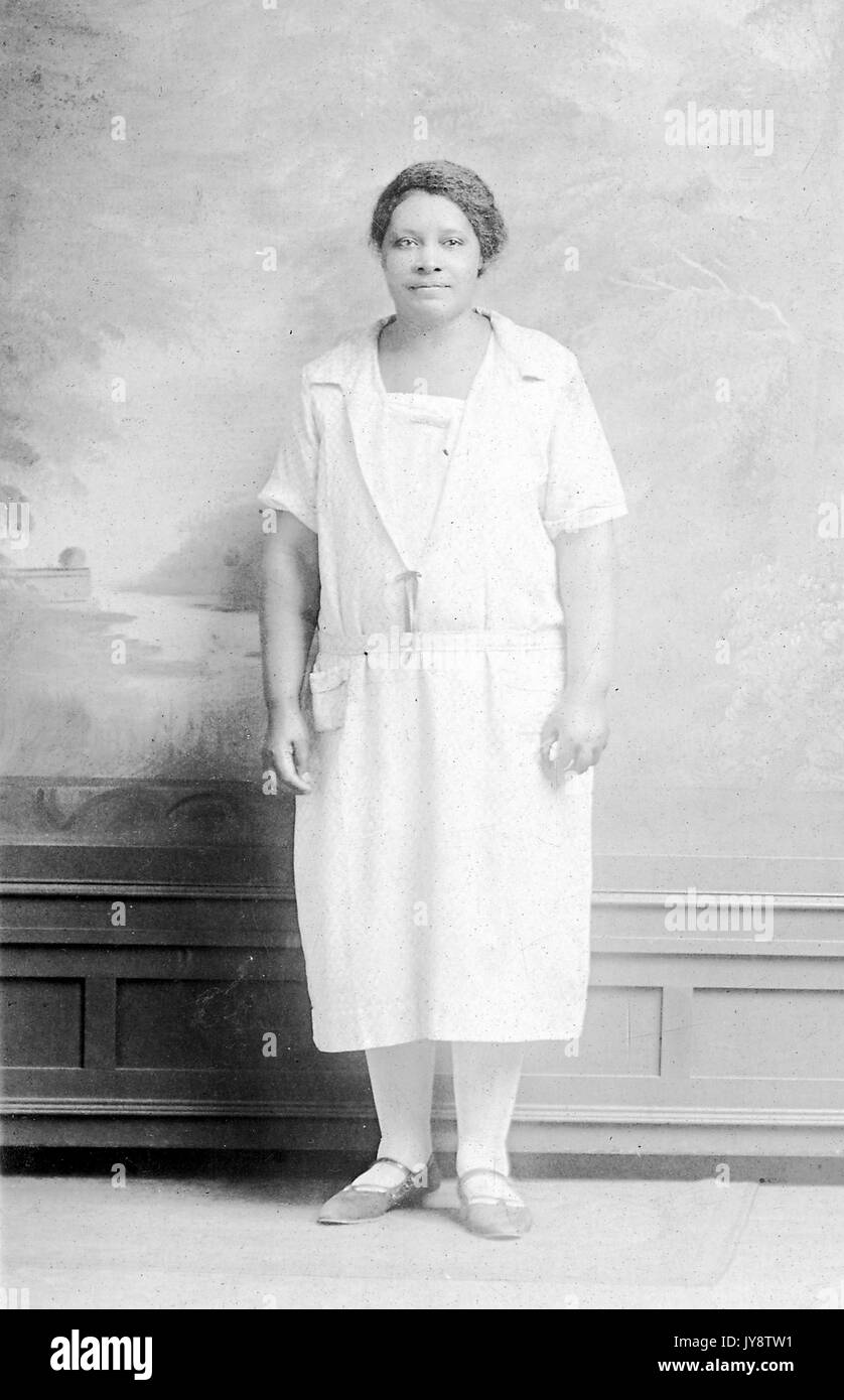 In voller Länge Porträt der jungen afrikanischen amerikanischen Frau in einem weißen Kleid und stehen vor einer gemalten Kulisse in einem Fotostudio, 1944. Stockfoto