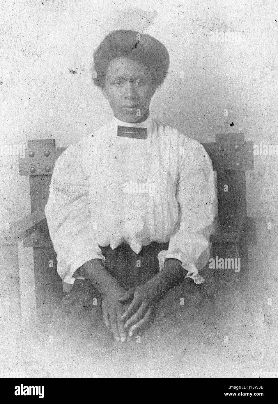Taille bis Portrait von Sitzen afrikanische amerikanische Frau, eine weiße Bluse und karierten Kleid, mit einem ernsten Gesichtsausdruck, die Hände auf dem Schoß, 1920 ruhte. Stockfoto